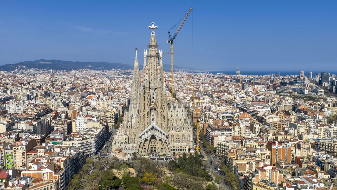 A large basilica standing clear of the surrounding rooftops in Barcelona, cranes still on its towers
