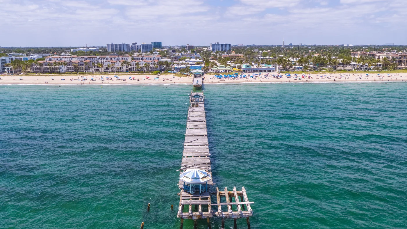 A fishing pier running out into green water with a crowded beach behind it