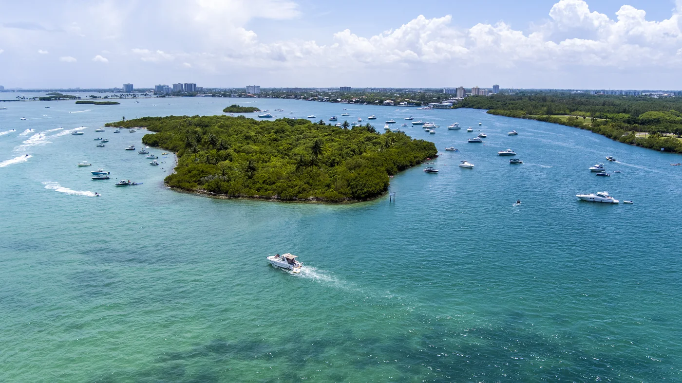 A mangrove island surrounded by anchored boats and crossing wakes in shallow turquoise water